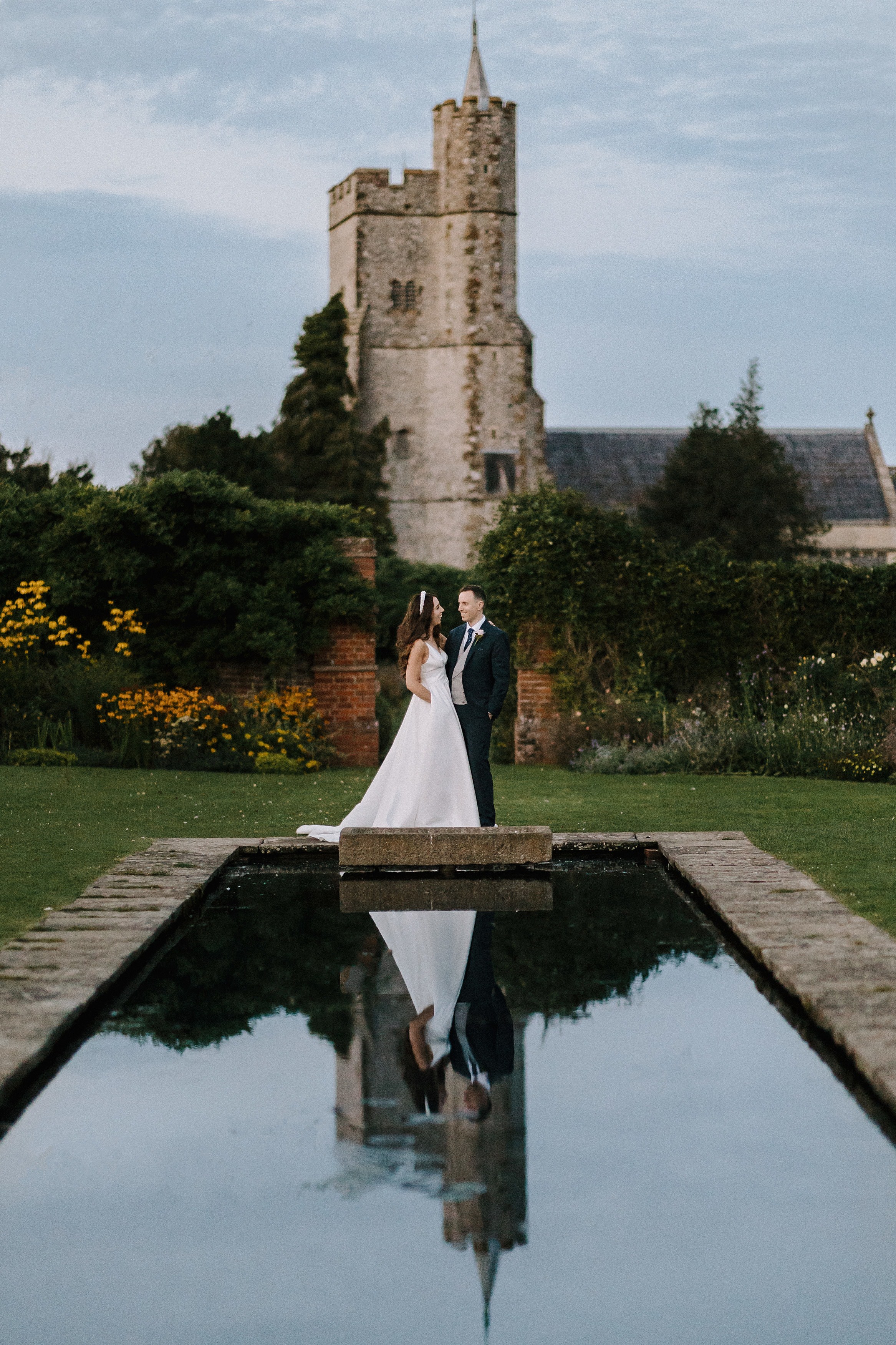Th bride and groom smiling whilst being reflected in the pond in the walled garden at Goodnestone Park with the Church of Holy Cross behind them